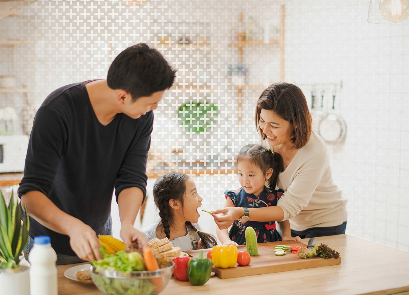 A family is gathered in a kitchen, with various family members interacting around a table. A toddler and a baby are present, along with adults who are wearing casual clothing. The scene includes a mixing bowl on the table and food items, set against an indoor backdrop with walls.