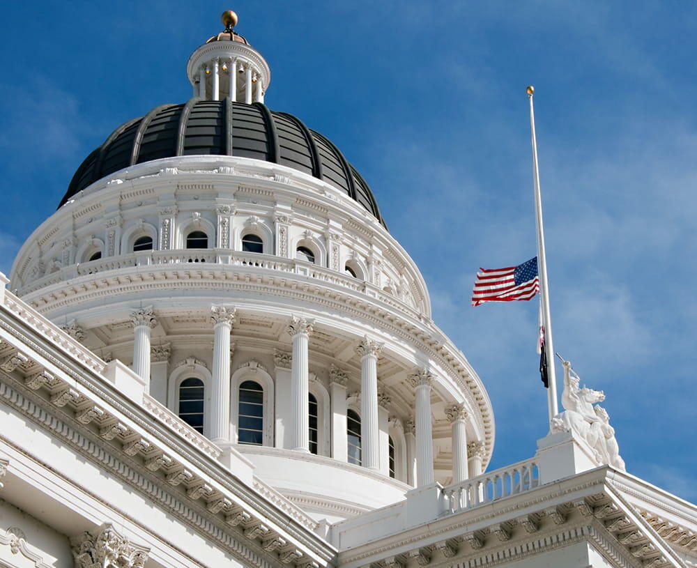 A flagpole stands atop a white building, with the California State Capitol visible in the background, under a blue sky dotted with clouds.